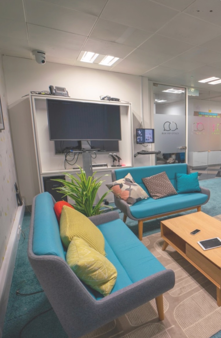 The John Lewis UX lab: two teal sofas and a coffee table arranged as a living room, facing a large monitor, recording equipment and a dome camera in the ceiling.