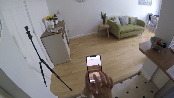 A second home: the participant points the phone at the empty floor beside a green sofa, with a second camera on a stand at the left of frame.