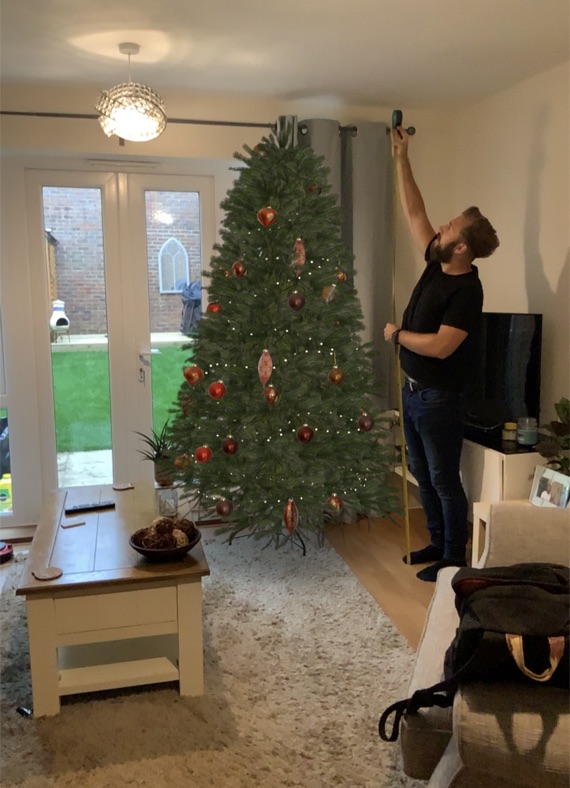 A living room with a decorated Christmas tree placed by augmented reality, while a person holds a tape measure up to the ceiling beside it.